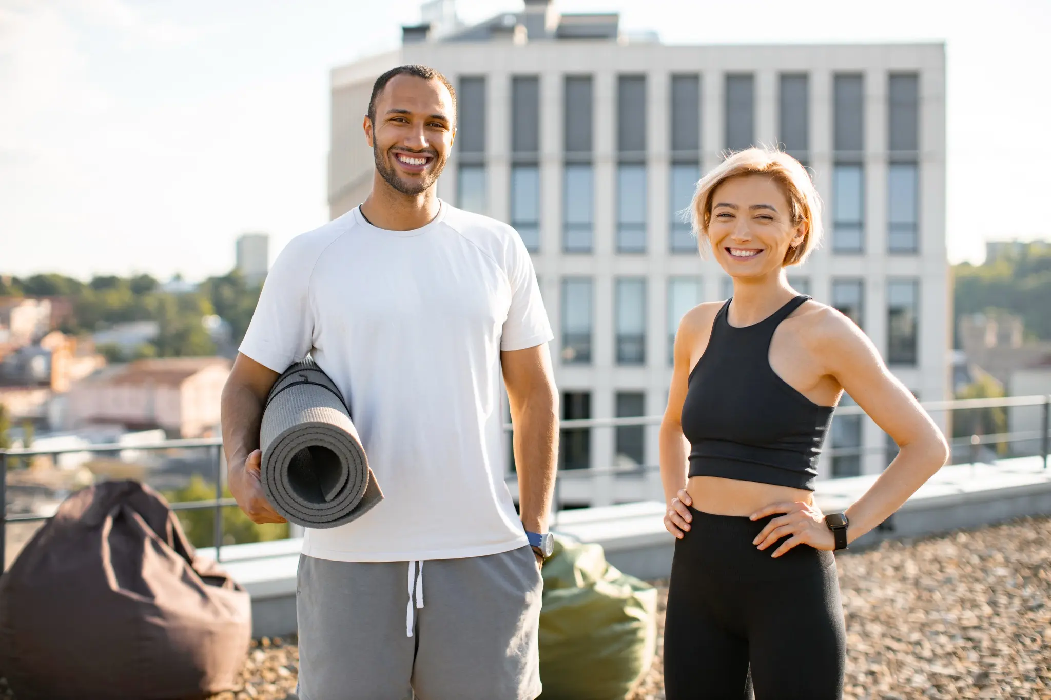 Ein Mann mit Yogamatte und eine Frau stehen lächelnd auf einer Dachterrasse.