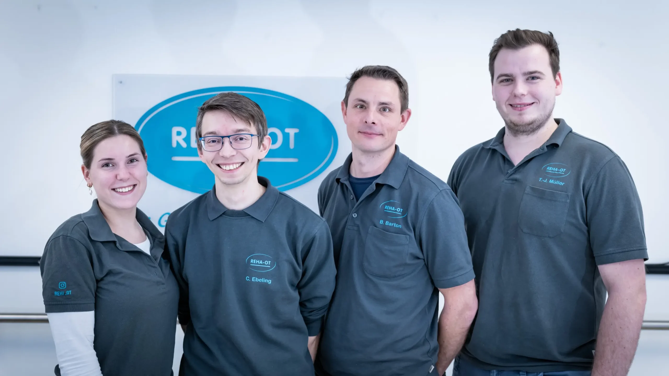 Vier lächelnde Personen in REHA-OT-Poloshirts vor einem Schild im Sanitätshaus Lüneburg.