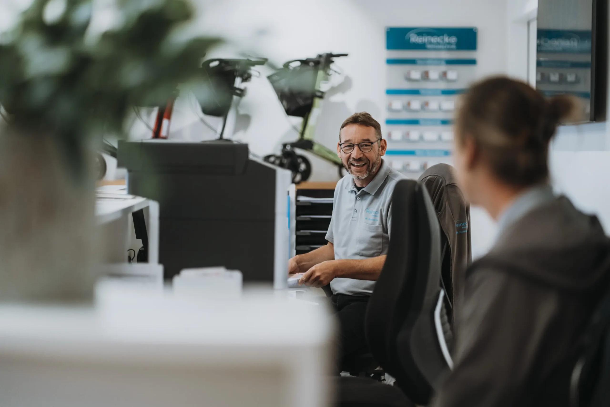 Zwei Personen in einem Büro, lächelnd im Gespräch. Hintergrund: Prospektständer, E-Scooter. Sanitätshaus.