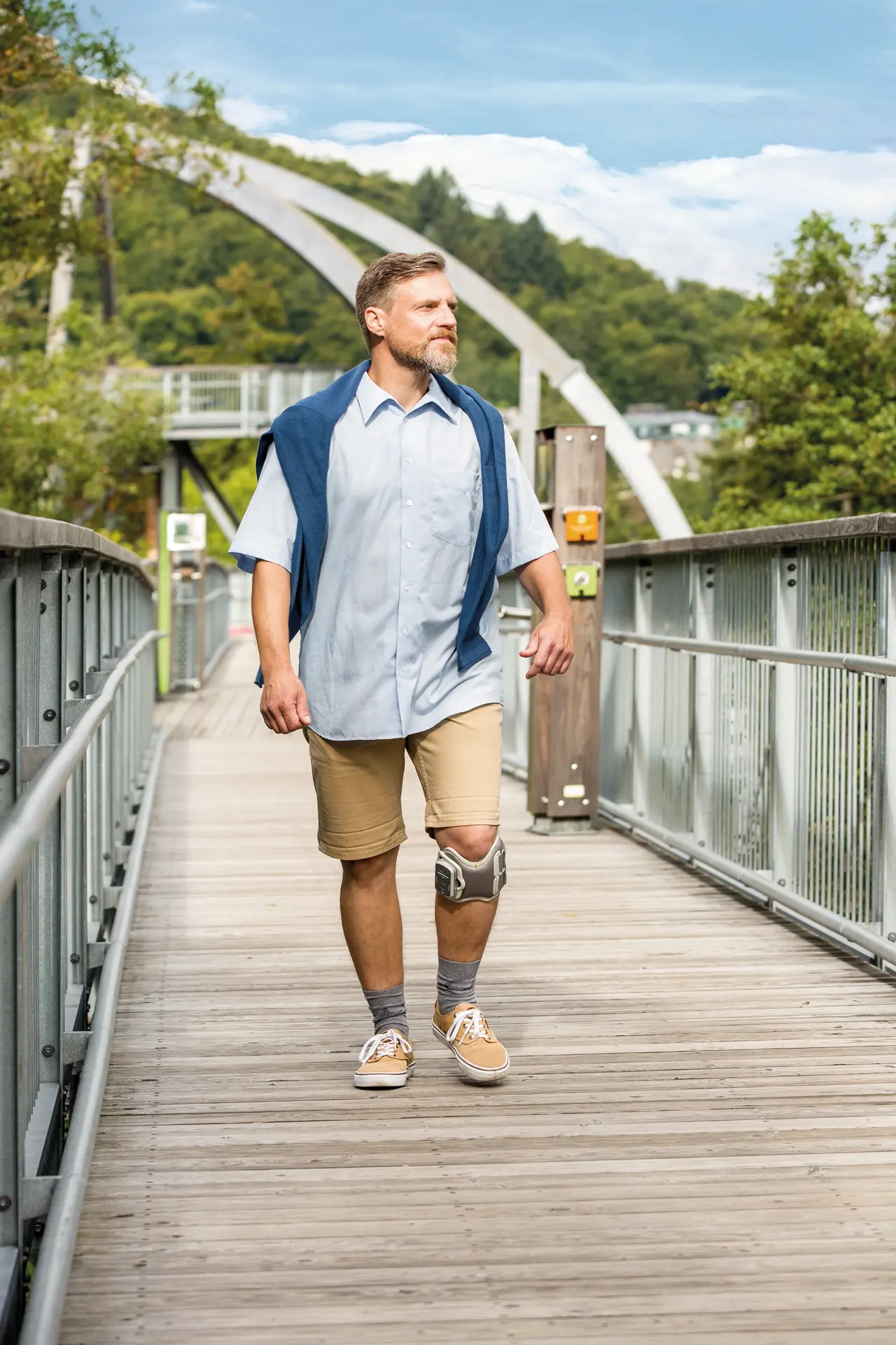 Ein Mann trägt eine Knieorthese und spaziert über eine Brücke in einer grünen Umgebung.
