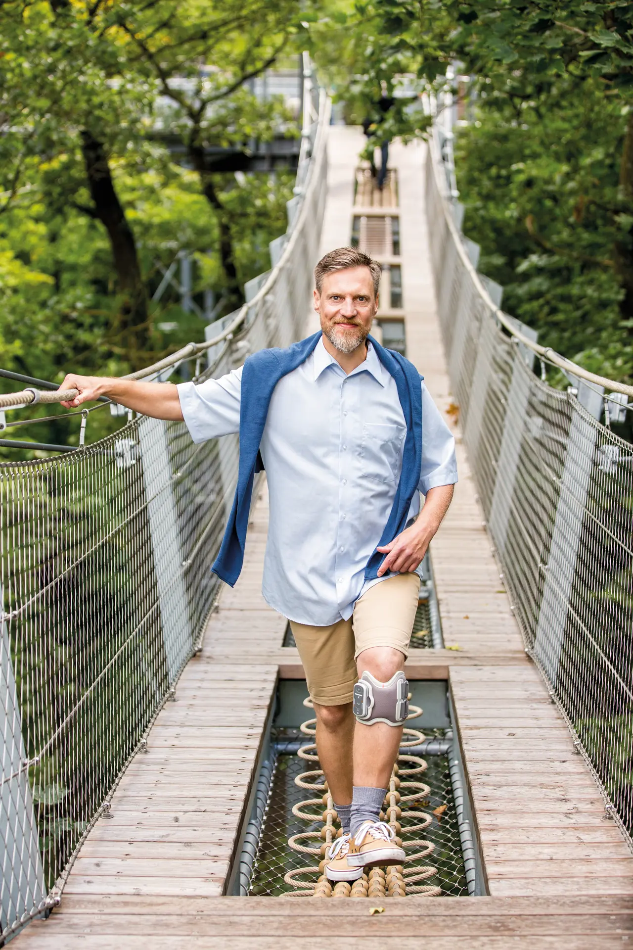 Ein Mann mit Knieorthese geht über eine Hängebrücke im Grünen.