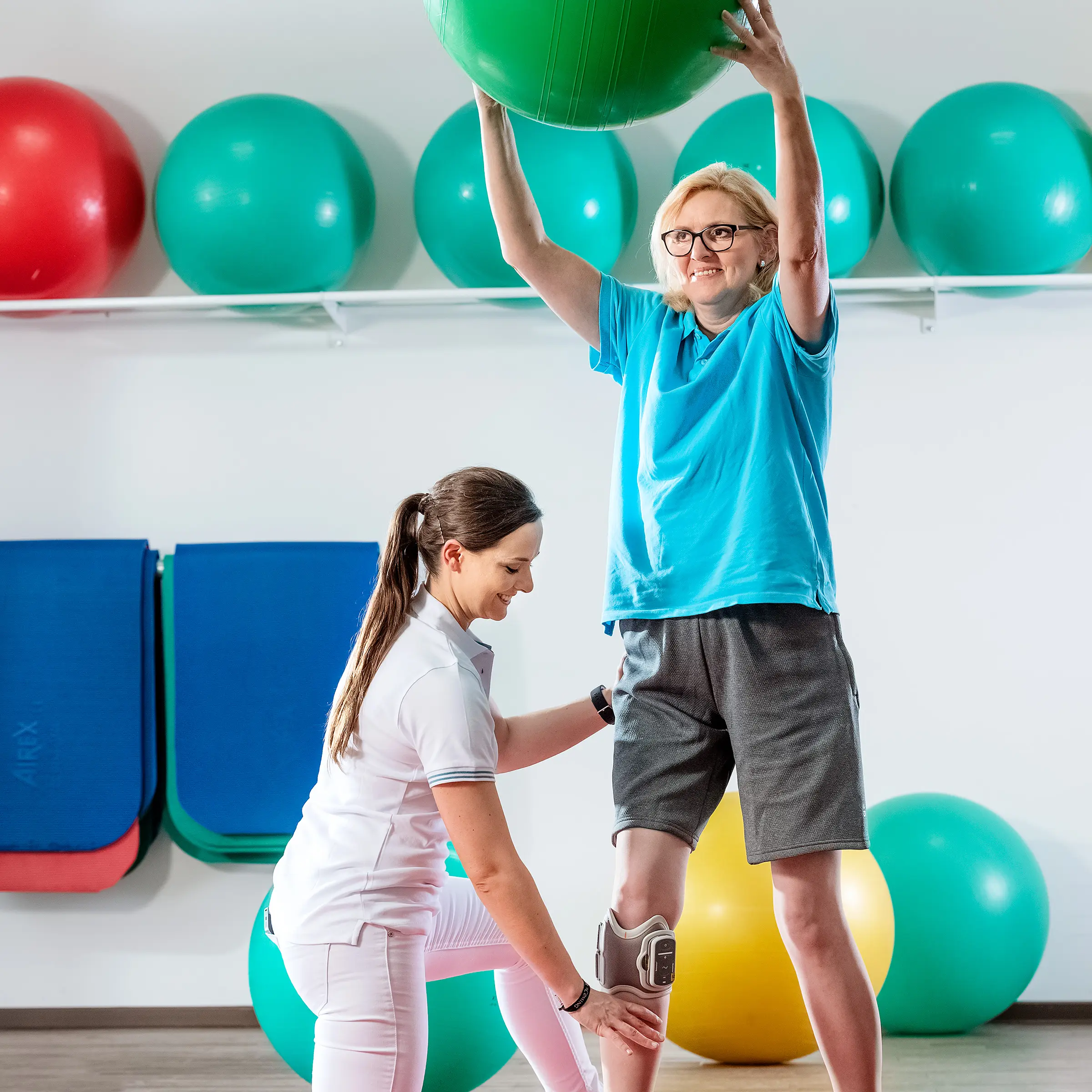 Eine Person mit Knieorthese trainiert mit Gymnastikball, unterstützt durch Physiotherapeutin.